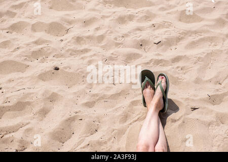 I piedi femminili con le gambe incrociate che indossa un paio di infradito unidentifed come noi guardiamo verso il basso su di una spiaggia di sabbia. Foto Stock