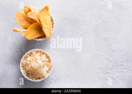 Disidratato ed essiccato di mango e scaglie di noce di cocco nella ciotola bianco su calcestruzzo sfondo luminoso. Vista da sopra. Foto Stock