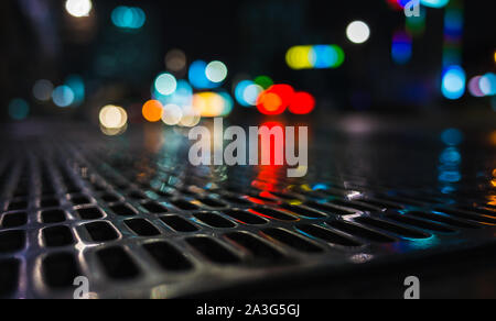 Abstract città notte foto con scintillante strada bagnata di fognature grattugiare sulla strada urbana e sfocate luci su uno sfondo, close-up foto con messa a fuoco selettiva Foto Stock