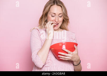 Giovane donna incinta indossando abiti rosa mangiando snack o dolci da big red coppa in plastica. Studio shot su sfondo rosa. Nutrizione durante la gravidanza Foto Stock