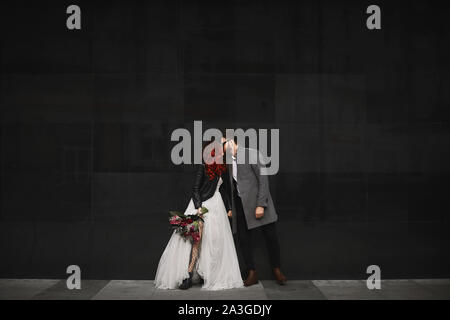 Bella coppia di appassionati baci all'aperto durante le cerimonie photoshoot. Slim giovane donna con i capelli rossi in eleganti giubbino in pelle e abito da sposa e Foto Stock