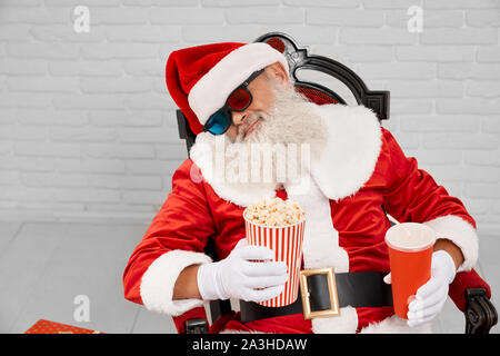 Stanco di natale tradizionale carattere in costume rosso dormire in una confortevole poltrona con popcorn e cola in mani. Coppia di Santa Claus prendendo nap prima di dare doni ai bambini felici. Foto Stock