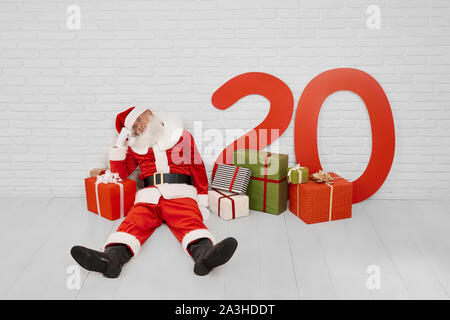 Stanco coppia Babbo Natale seduto sul pavimento bianco vicino al red big box regalo con i numeri di carta di 2020 Anno Nuovo. Il tempo di Natale, buone feste e tradizioni. Foto Stock