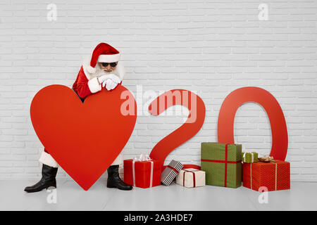 Sorridente San Nicola in costume rosso, bianco guanti e occhiali neri appoggiarsi sul grande carta rossa cuore con sullo sfondo di un muro bianco. Concetto di Nuovo Anno 2020 Foto Stock