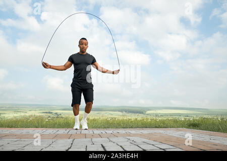 Attivo, bello, concentrata atleta jumping con salto con la corda all'aperto, guardando verso il basso. Uomo Forte con corpo sportivo in nero di t-shirt e shorts formazione, facendo esercizi. Foto Stock