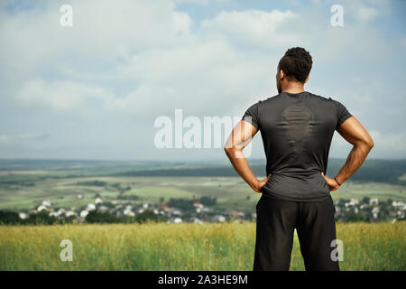 Vista posteriore del muscolare sportivo africani che indossa in nero di t-shirt e pantaloncini neri. Bello, forte, atletico uomo in piedi e in posa all'aperto. Atleta tenendo le mani sui fianchi. Foto Stock