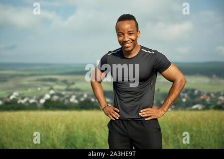 Ritratto di sorridere, bello sportivo all aperto con le mani sui fianchi. Muscoloso atleta indossa in nero di t-shirt e shorts. Atleta positivo in posa sullo sfondo della natura e il blu del cielo. Foto Stock