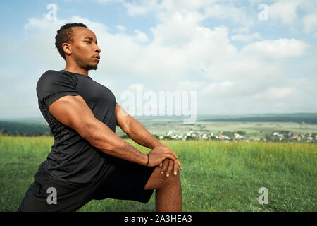 Bello l'uomo africano facendo esercizi in mattina all'esterno. Sportivo con corpo muscoloso indossa in nero di t-shirt e shorts stretching. Atleta in posa sullo sfondo del cielo blu. Foto Stock