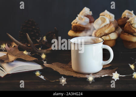 Caldo e fumante tazza di caffè con panini alla cannella e libro aperto il fuoco selettivo sulla bevanda con estrema sfocato in primo piano e sullo sfondo. Foto Stock