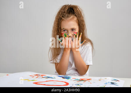 Bambina in t-shirt bianca con seduta a tavola con carta da filtro Whatman e vernici su di esso, posa con dipinto il viso e le mani. Isolato su bianco. Medie di close-up. Foto Stock