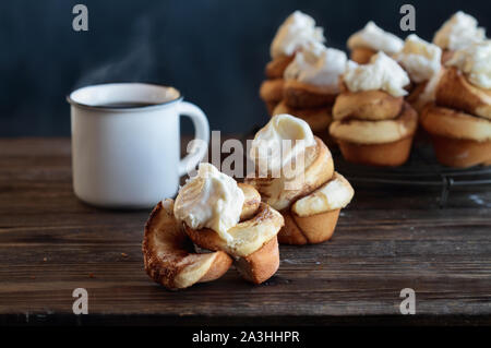 Fresche fatte in casa tall cinnamon roll con glassa di formaggio cremoso. Fumante tazza di caffè in background con più panini raffreddamento su un fornaio rack. Foto Stock