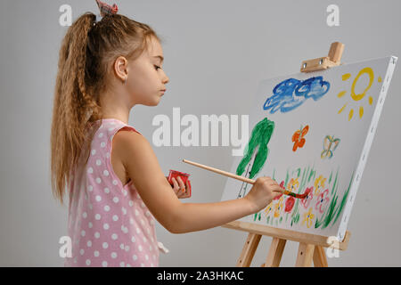 Bambina artista in un abito rosa è in piedi dietro il cavalletto e pittura con pennello su tela, isolato su bianco di sfondo per studio. Medie di close-up. Foto Stock