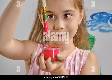 Bambina artista in un abito rosa è in piedi dietro il cavalletto e pittura con pennello su tela, isolato su bianco di sfondo per studio. Medie di close-up. Foto Stock