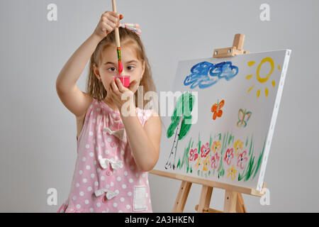 Bambina artista in un abito rosa è in piedi dietro il cavalletto e pittura con pennello su tela, isolato su bianco di sfondo per studio. Medie di close-up. Foto Stock