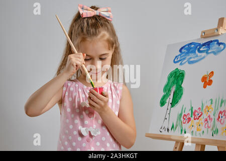 Bambina artista in un abito rosa è in piedi dietro il cavalletto e pittura con pennello su tela, isolato su bianco di sfondo per studio. Medie di close-up. Foto Stock