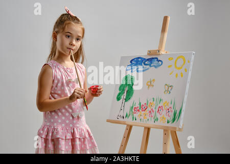 Bambina artista in un abito rosa è in piedi dietro il cavalletto e pittura con pennello su tela, isolato su bianco di sfondo per studio. Medie di close-up. Foto Stock