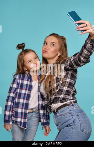 Mamma e figlia vestito in camicie a scacchi e blue jeans denim stanno rendendo selfie mentre posa contro un blu di sfondo per studio. Close-up shot. Foto Stock