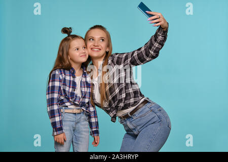 Mamma e figlia vestito in camicie a scacchi e blue jeans denim stanno rendendo selfie mentre posa contro un blu di sfondo per studio. Close-up shot. Foto Stock