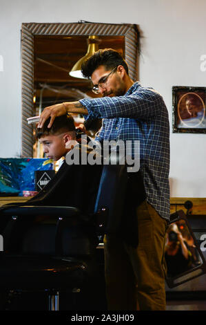 Un ragazzo con grembiule siede nel Barbiere sedia mentre avente taglio di capelli alla moda nel quartiere alla moda di '81 Barbieri shop in Tucson, AZ Foto Stock