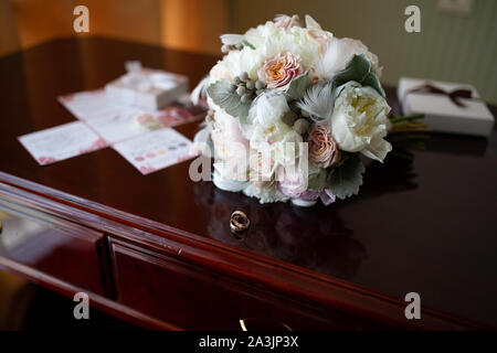 Wedding bouquet di peonie e rose giacente su un vintage in legno tavolo vicino gli anelli di nozze sullo sfondo del bando negli appartamenti di prepa Foto Stock