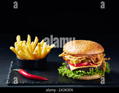 Cheeseburger con polpetta di carne, formaggio cheddar, croccante di cipolle fritte, lattuga, pomodori a fette cetrioli sottaceto su una pietra nera bordo con patate fritte Foto Stock