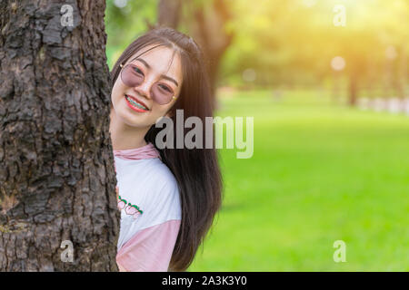 Felice asiatici bretelle dentali ragazza indossando occhiali da sole felice nascondino a tree park ritratto all'aperto. Foto Stock