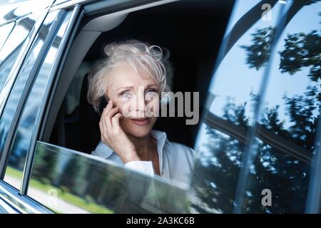 Allegra donna matura in business suit seduti sul sedile posteriore della sua automobile e parlando al telefono cellulare. Senior imprenditrice usando il telefono mentre si è in viaggio da Foto Stock