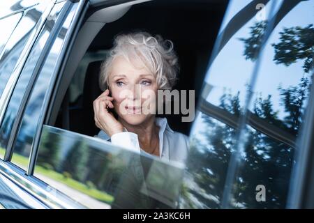 Allegra donna matura in business suit seduti sul sedile posteriore della sua automobile e parlando al telefono cellulare. Senior imprenditrice usando il telefono mentre si è in viaggio da Foto Stock