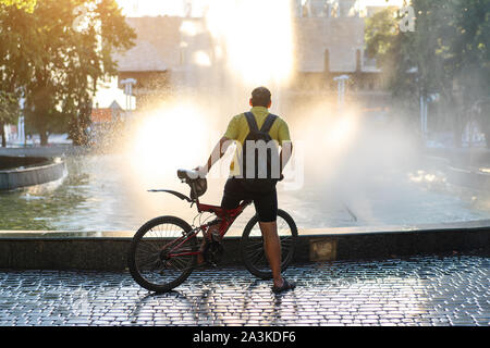 Irriconoscibile ciclista sorge vicino alla fontana tenendo in bicicletta in mani su un luminoso giorno d'estate. Estate le persone attive. Uno stile di vita sano Foto Stock