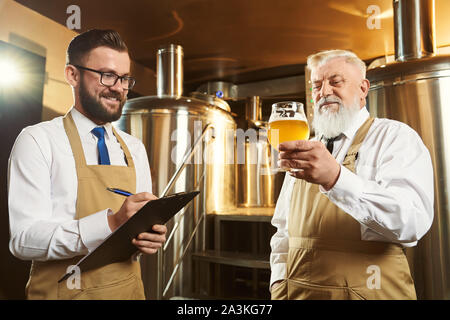 Produzione di birra. Due maschi lavoratori birreria esaminando la birra. Anziani brewer tenendo il vetro con la birra, prove. Uomo bello tenendo la cartella e penna, sorridente e la scrittura. Foto Stock