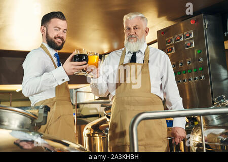 Due produttori di birra in piedi in mini birreria vicino ad apparecchiature, tenendo i bicchieri con la luce e la birra scura, cercando e sorridente. I lavoratori che indossa in camice bianco e marrone e grembiuli. Concetto di birra brewering processo. Foto Stock