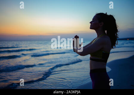 Silhouette di giovane donna in abiti fitness sulla Ocean Shore in serata meditando. Foto Stock