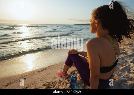 Visto da dietro la donna attiva nello sport vestiti con bottiglia di acqua sedersi e rilassarsi dopo allenamento in riva al mare in serata. Foto Stock