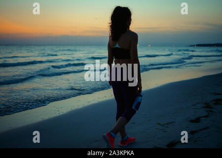 Visto da dietro sport fitness donna in abiti di fitness con una bottiglia di acqua minerale a piedi dopo allenamento sulla Ocean Shore in serata. Foto Stock