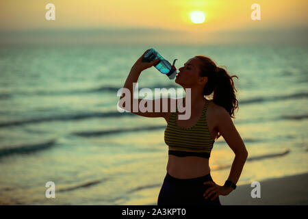 Silhouette di montare la donna in stile sport vestiti in spiaggia al tramonto bevendo acqua dalla bottiglia. Foto Stock