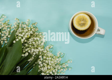 Vista superiore della tazza di tè con la fetta di limone e il giglio della valle dei fiori sulla pastello blu sullo sfondo. Copia dello spazio. Mockup per la progettazione. Foto Stock