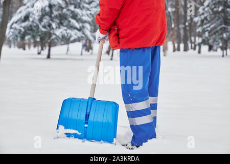 Vista ritagliata della bidello maschio nel processo di lavorazione in inverno nel parco. Uomo in uniforme blu e giacca rossa rimozione neve da marciapiedi con pala. Concetto di manutenzione e servizio. Foto Stock