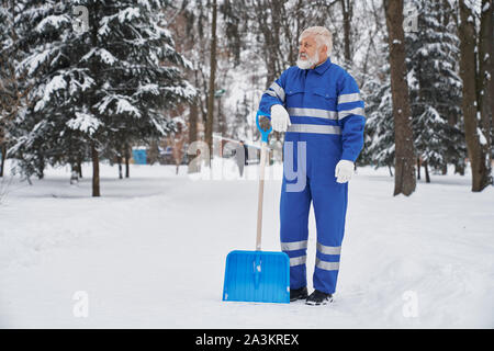 Bidello maschio in uniforme blu in piedi in posizione di parcheggio e il riposo durante la pulizia dei marciapiedi da neve. Lavoratore di sesso maschile mantenere attrezzature e cercando riposo in inverno. Concetto di compensazione e di servizio. Foto Stock