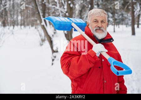 Vista frontale di un uomo guarda la fotocamera e sorridere mentre il parco di pulizia dalla neve in inverno. Lavoratore di sesso maschile in giacca rossa mantenendo la pala sulla spalla e posa. Concetto di inverno e cancellazione. Foto Stock