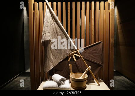 Sauna tradizionale stuff - una bandiera, un ventilatore, asciugamani e un secchio con un cucchiaio di legno su un banco di lavoro con doccia con sauna in background Foto Stock