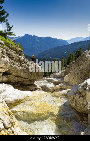 Giovane donna attraente relax su una roccia sul lato di un piccolo ruscello di montagna in un pittoresco paesaggio di montagna nelle Dolomiti in Italia Foto Stock
