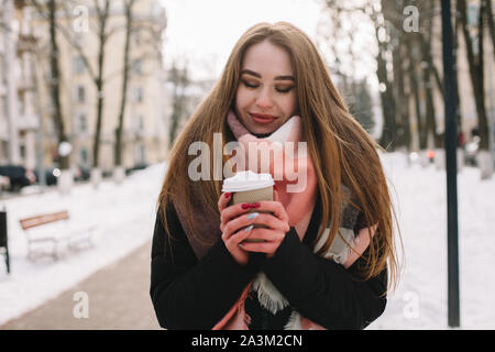 Felice giovane donna con tazza monouso indossando vestiti caldi mentre si cammina sulla strada di città durante la stagione invernale Foto Stock