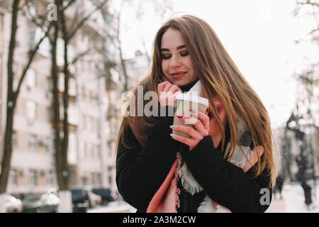 Felice giovane donna con tazza monouso indossando vestiti caldi mentre si cammina sulla strada di città durante la stagione invernale Foto Stock