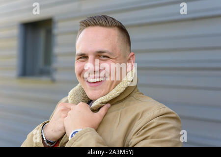 Felice giovane con un sorriso carismatico in piedi all'aperto in un freddo giorno di una calda giacca di pelle di pecora passando per grin presso la fotocamera Foto Stock