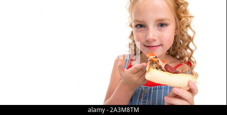 Una bambina con heap rosso i capelli in una maglia rossa e blu e bianco tuta in una striscia mangia una grande pizza. Foto Stock