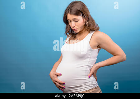 Giovane donna incinta che indossa una camicia bianca tenendo il suo ventre mantenendo una mano sulla sua schiena soffre di mal di schiena. Studio shot su sfondo blu Foto Stock