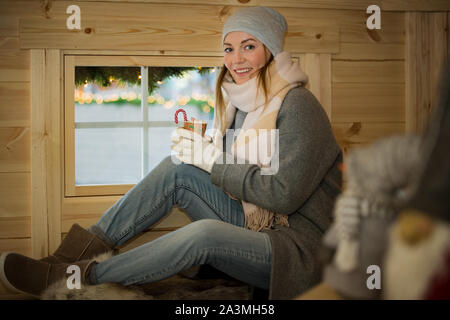 Giovane donna bere tazza di cioccolata calda con marshmallow maglia da indossare caldo cappello e sciarpa. Seduta accanto alla finestra in finlandese baita in legno Foto Stock