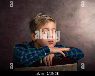 Splendido piccolo ragazzo biondo, triste, poggiando su una sedia Foto Stock
