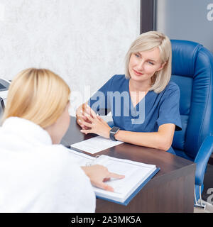 Servizio di cosmetologia. Giovane donna alla clinica di bellezza seduta elenco lettura delle procedure per discutere con il medico positivo Foto Stock