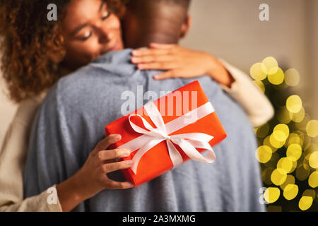 Amorevole donna afro abbracciando il marito, tenendo regalo di natale Foto Stock